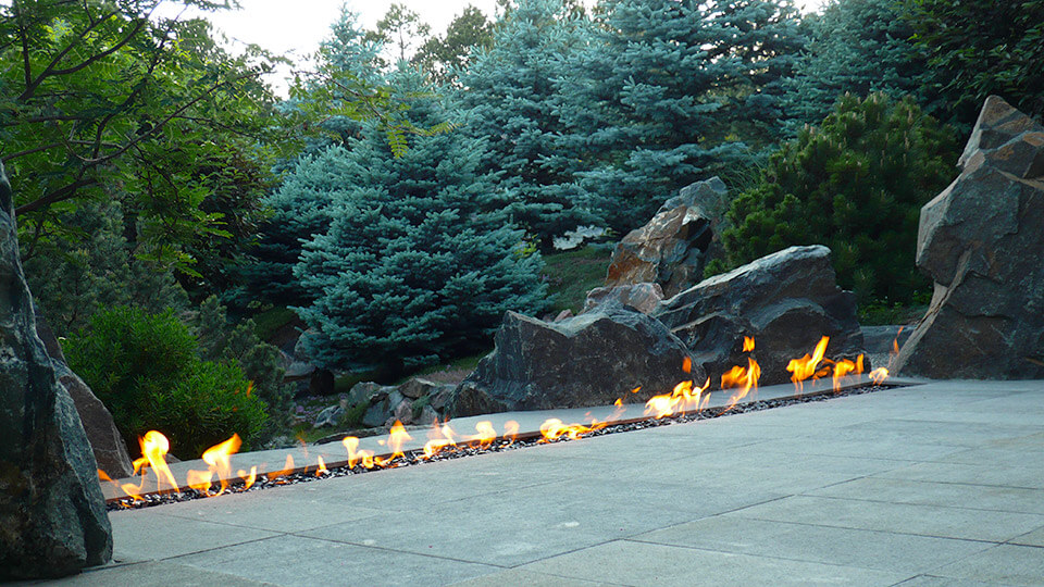 Rectangular Row fire fit in backyard with stones, plants and trees