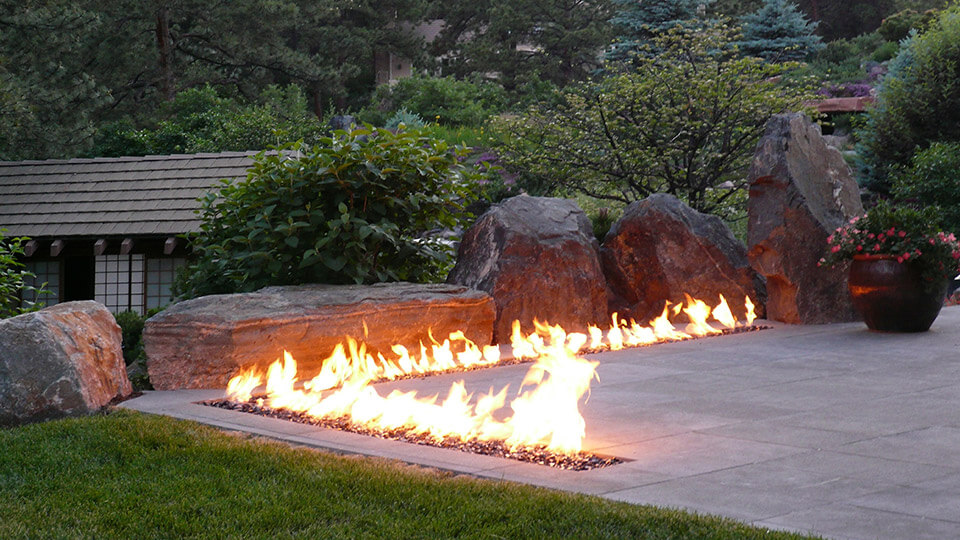 L-shaped fire pit in backyard with stones, plants and trees