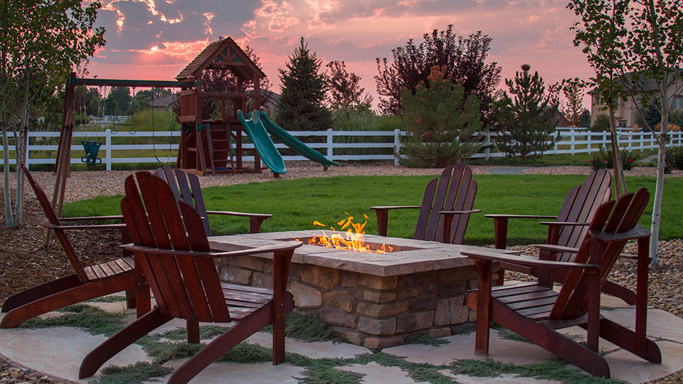 Fire pit surrounded by chairs along with kids playground and double swing in the backyard with sky view