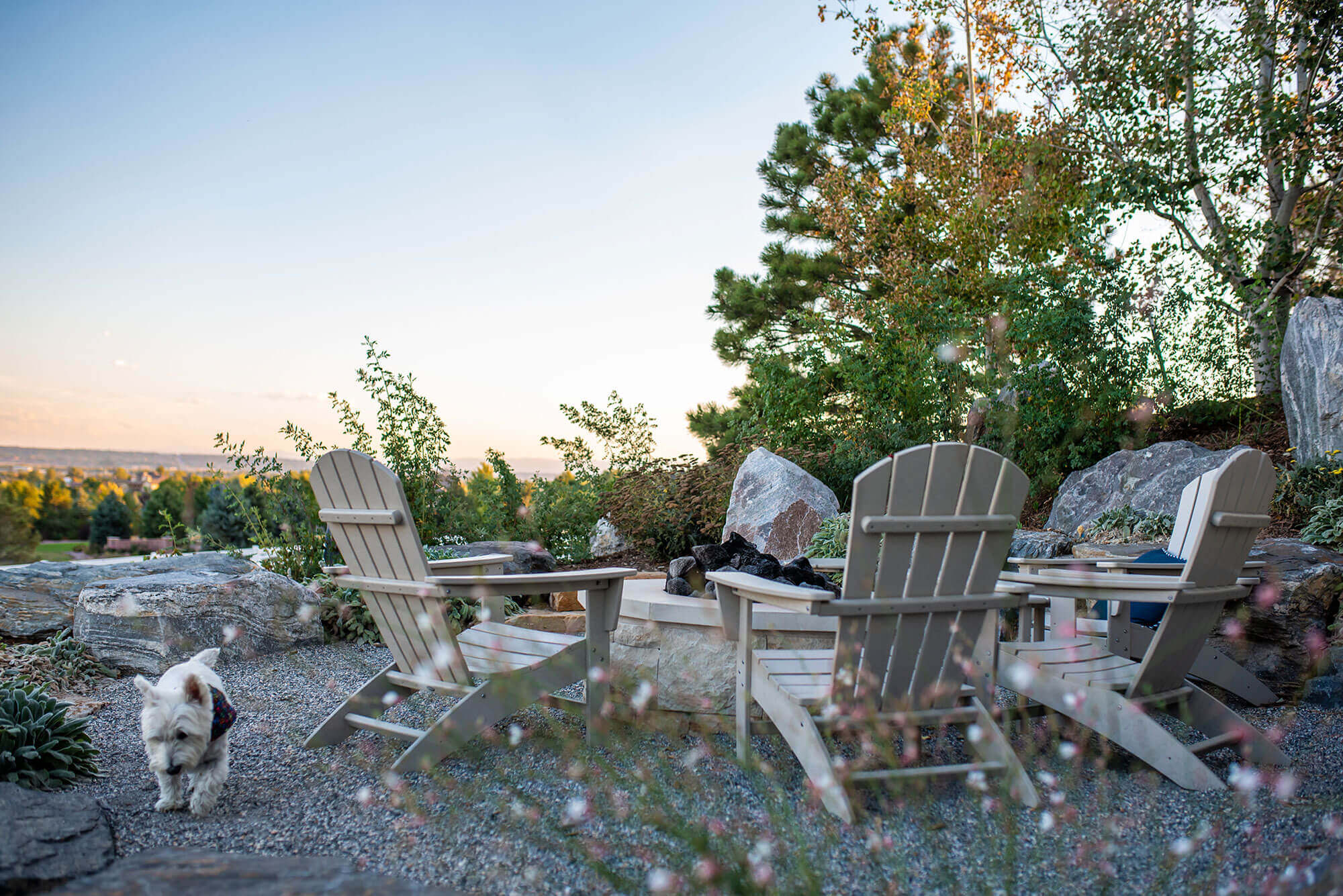 Fire pit area filled with rocks along with some chairs, fire pit, plants and trees