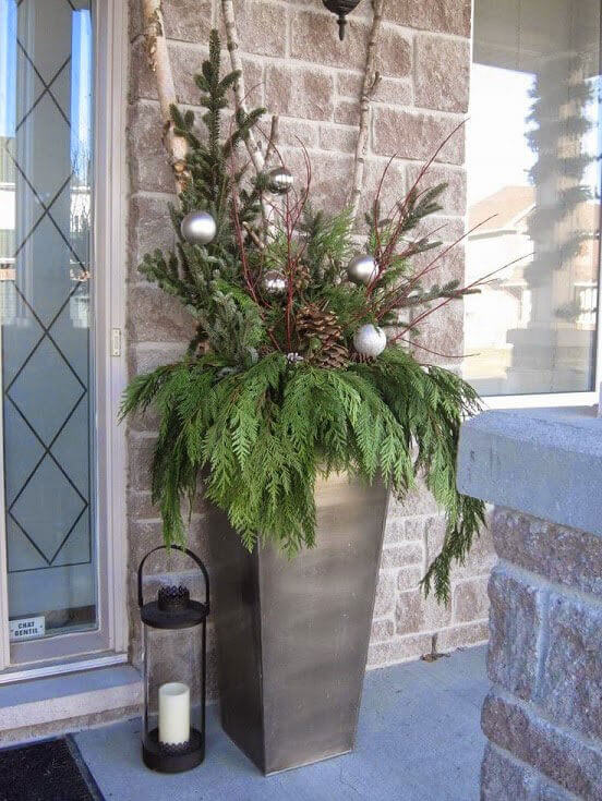 A plant is decorated with christmas bulbs, ornaments and a candle side of it in front of the house