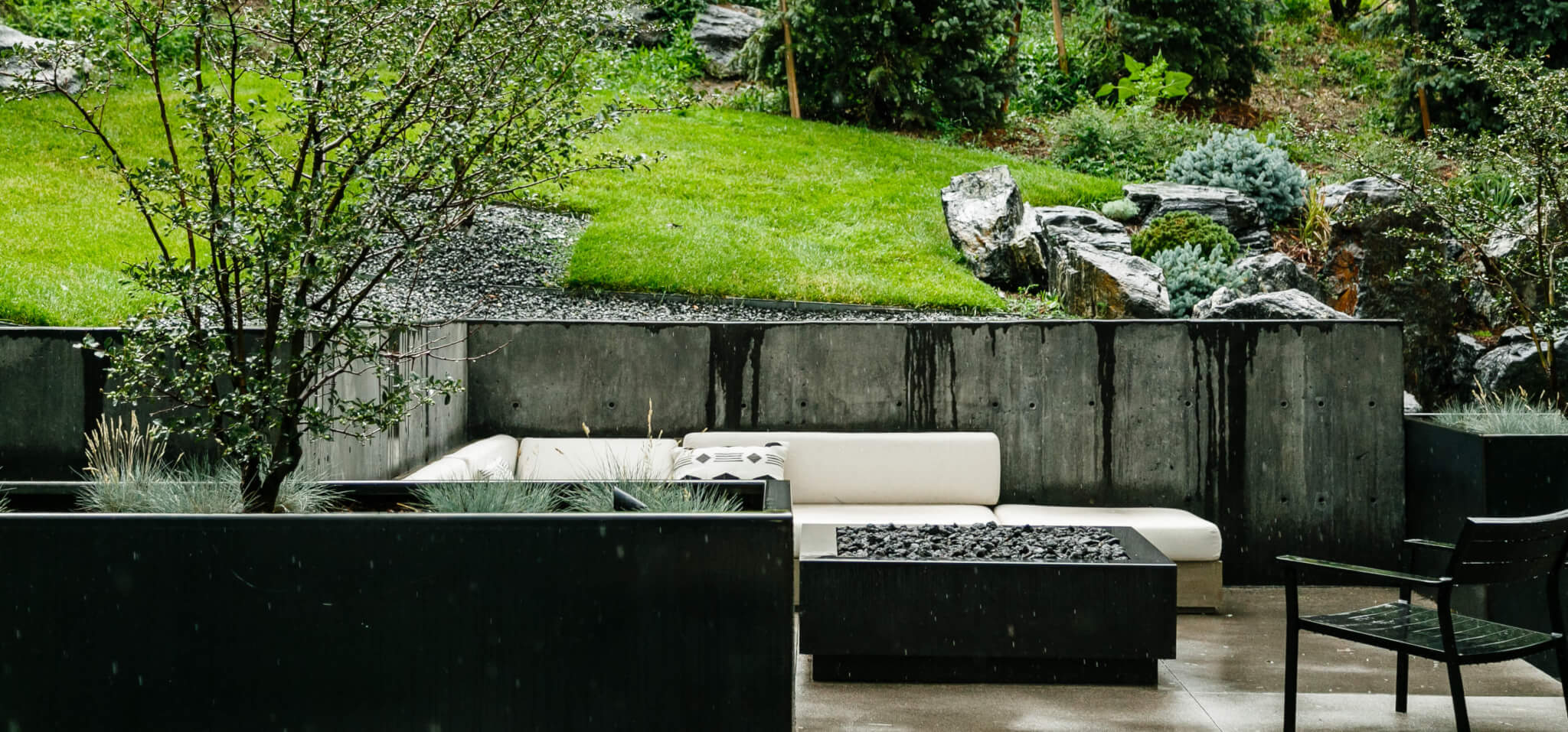 Sitting area with fire pit, sofa, stone planter filled with grass plant and tree