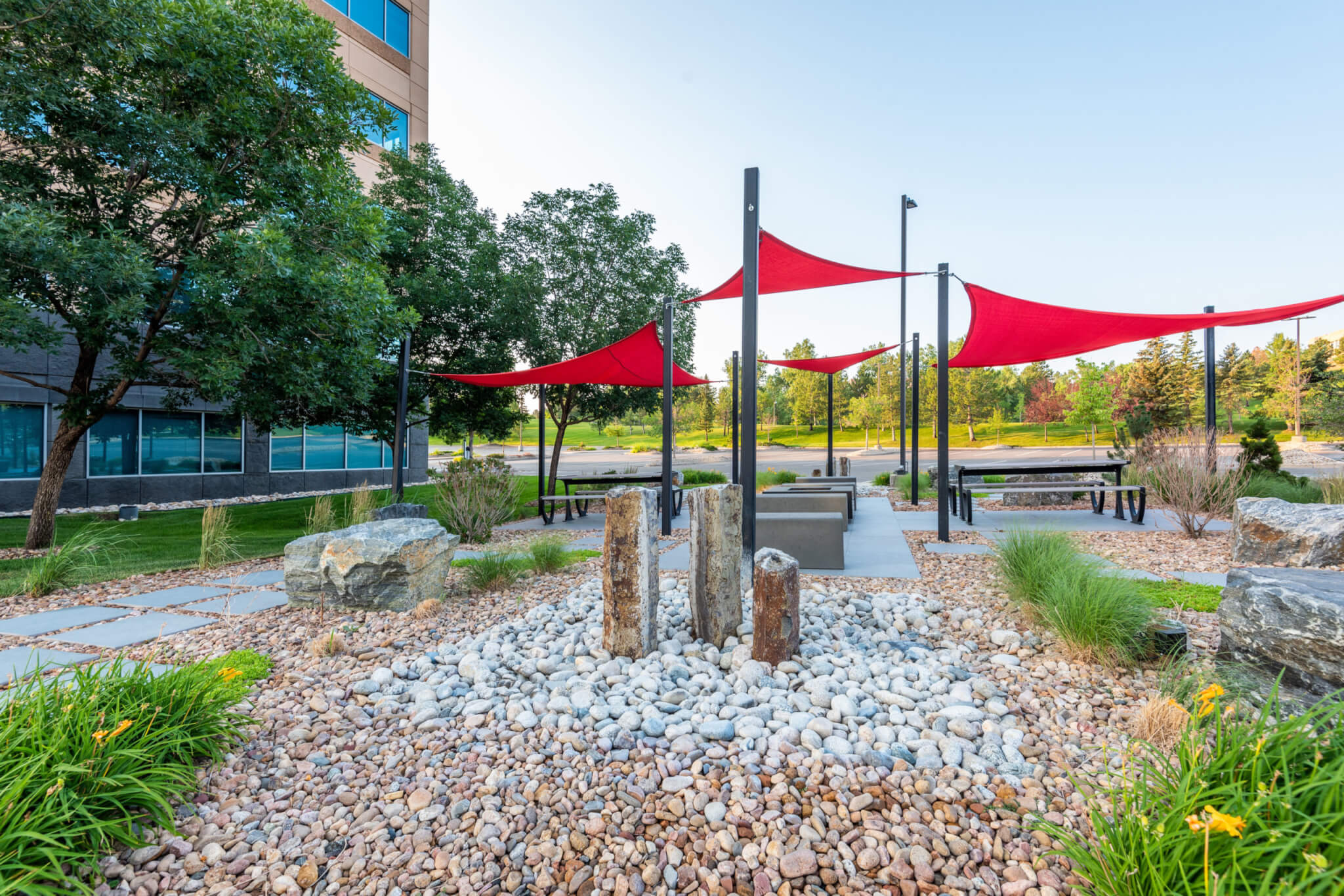 Sitting area filled with rock sitting benches, bushes, and trees along with red fabric tensile membrane with sail shade structure