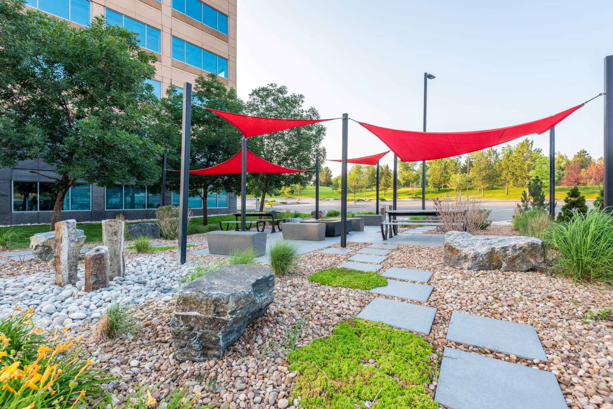 Sitting area filled with rocks, stones, sitting benches along with red fabric tensile membrane with sail shade structure in front of the building