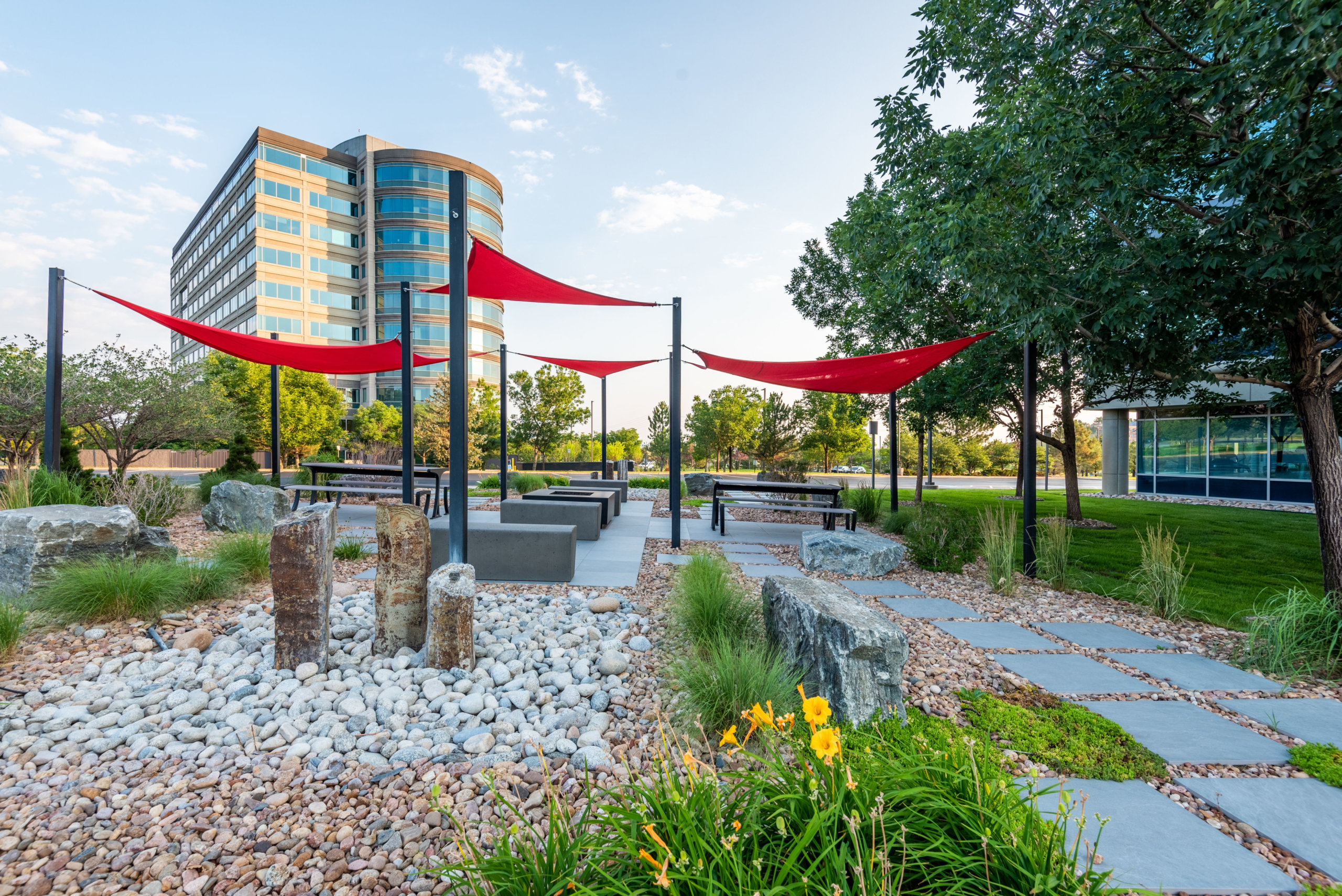 Sitting area filled with rocks, stones, sitting benches along with red fabric tensile membrane with sail shade structure in front of the building