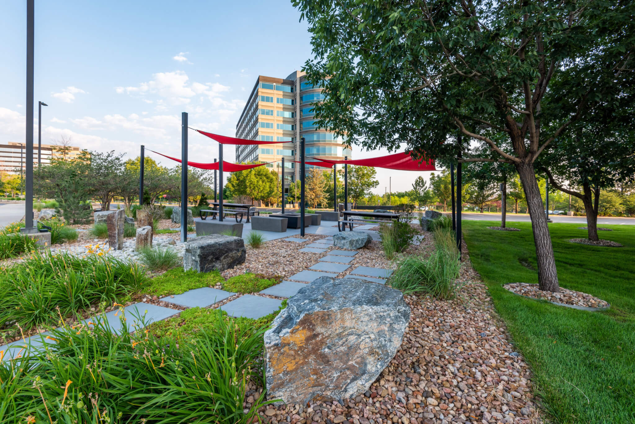 Sitting area filled with rocks, stones, trees, sitting benches along with red fabric tensile membrane with sail shade structure in front of the building