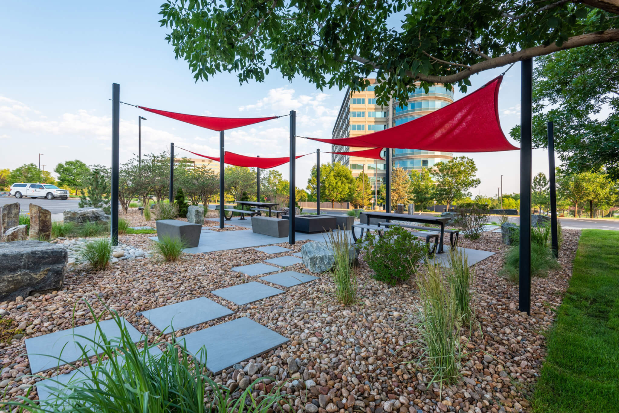 Sitting area filled with rocks, stones, grass plants, sitting benches along with red fabric tensile membrane with sail shade structure in front of the building