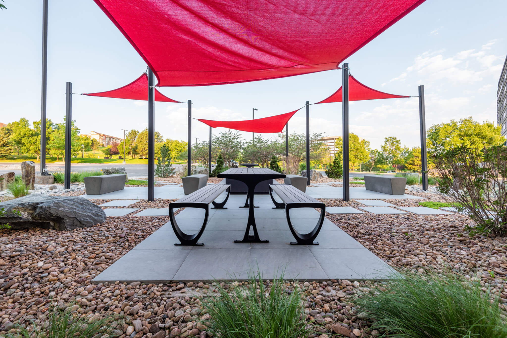 Sitting area filled with stone benches, bushes, and trees along with red fabric tensile membrane with sail shade structure