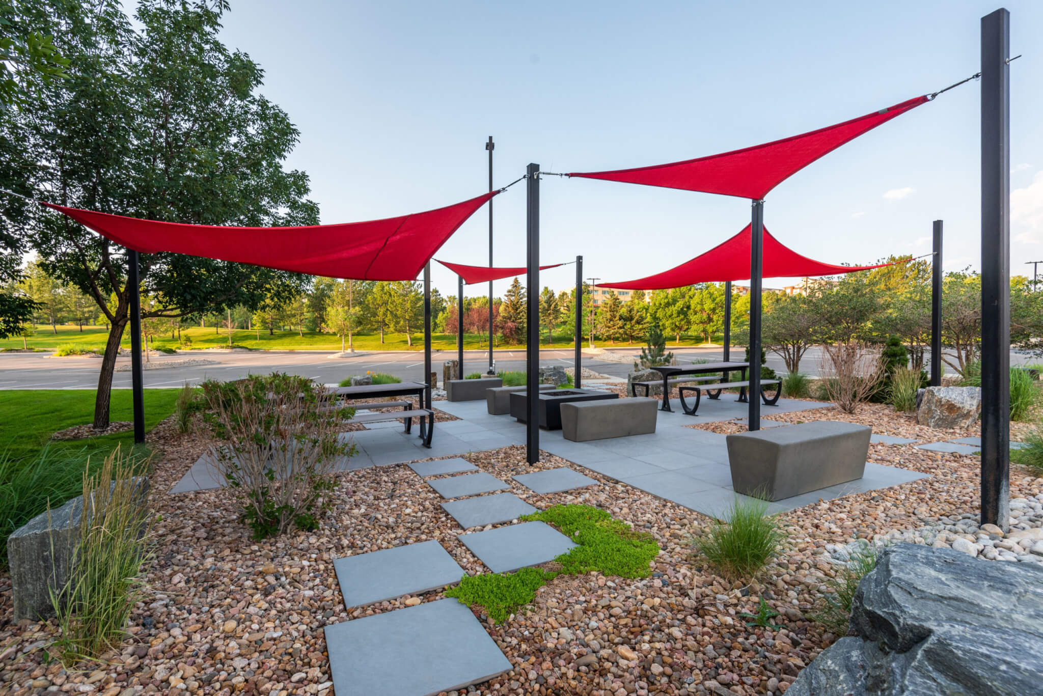 Sitting area filled with stone sitting benches, bushes, and trees along with red fabric tensile membrane with sail shade structure