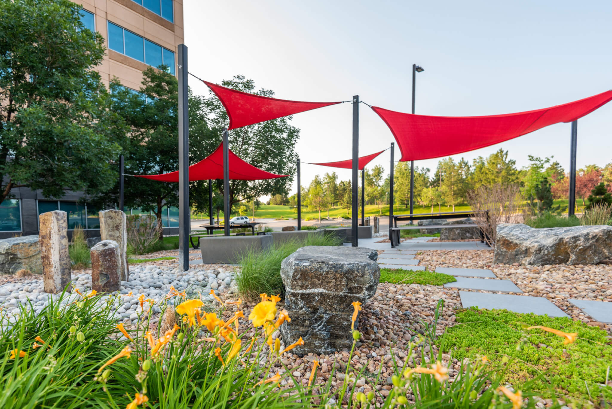 Sitting area filled with rock sitting benches, bushes, and trees along with red fabric tensile membrane with sail shade structure