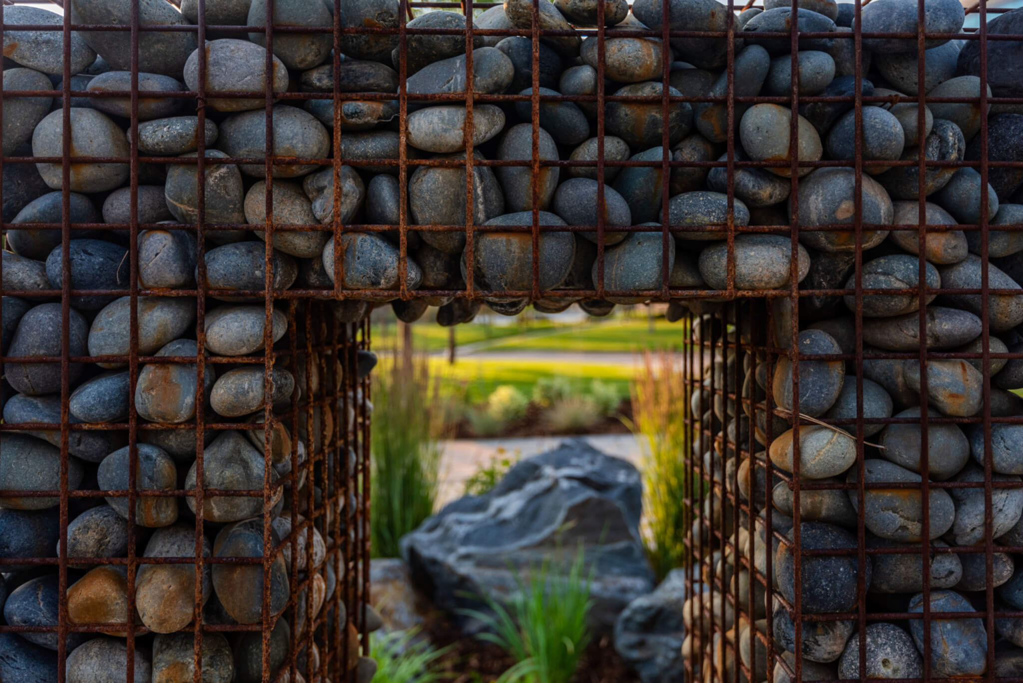 A metal cage filled with rocks in a sitting area with grass plants and trees