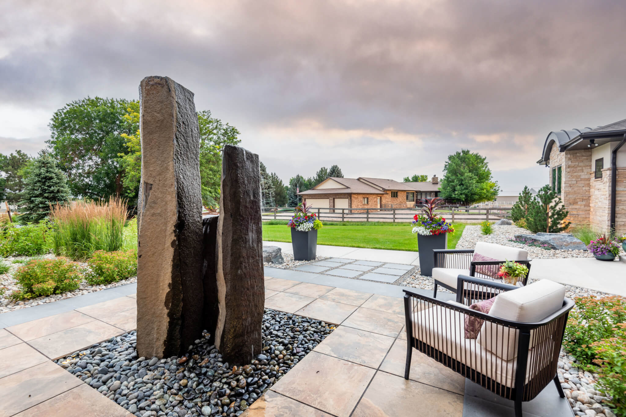 Rock sculpture in garden area with flower plant pots, chairs, grass plants and trees