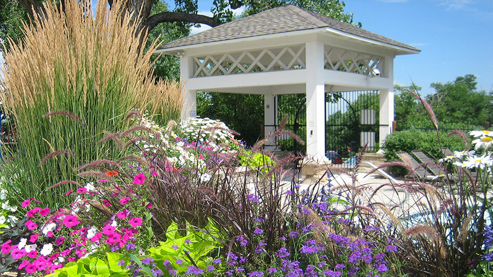 There is a gazebo in the garden filled with flower plants and trees