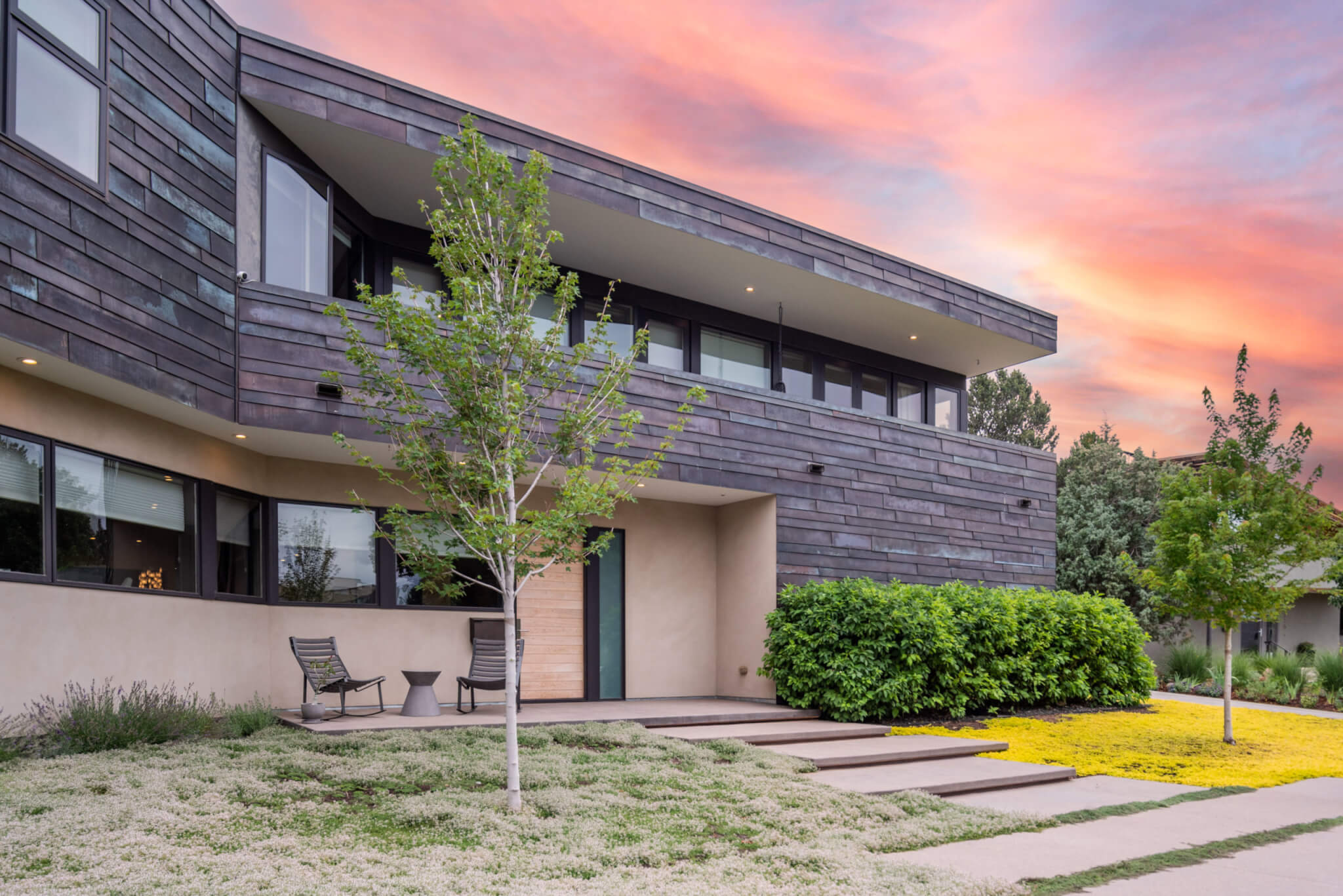 Front view of a house with tinted glass windows, some chairs, plants and trees