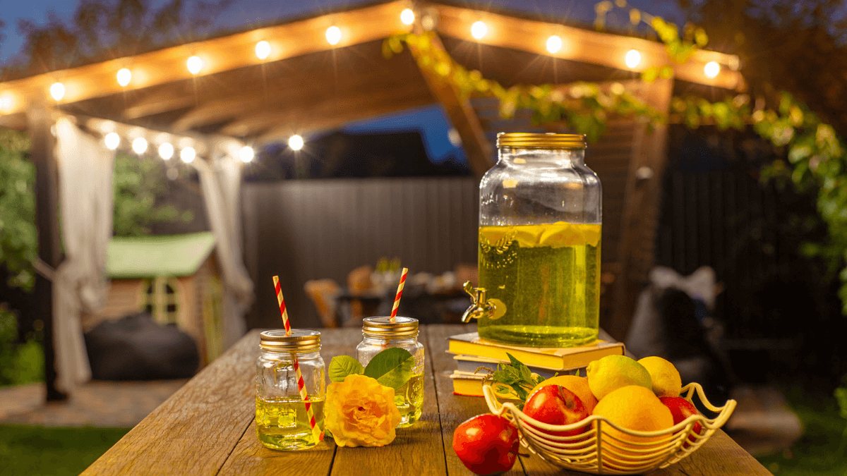 backyard at night light up by bistro string lights. Lemonade present in the foreground with fruit and flowers on the side