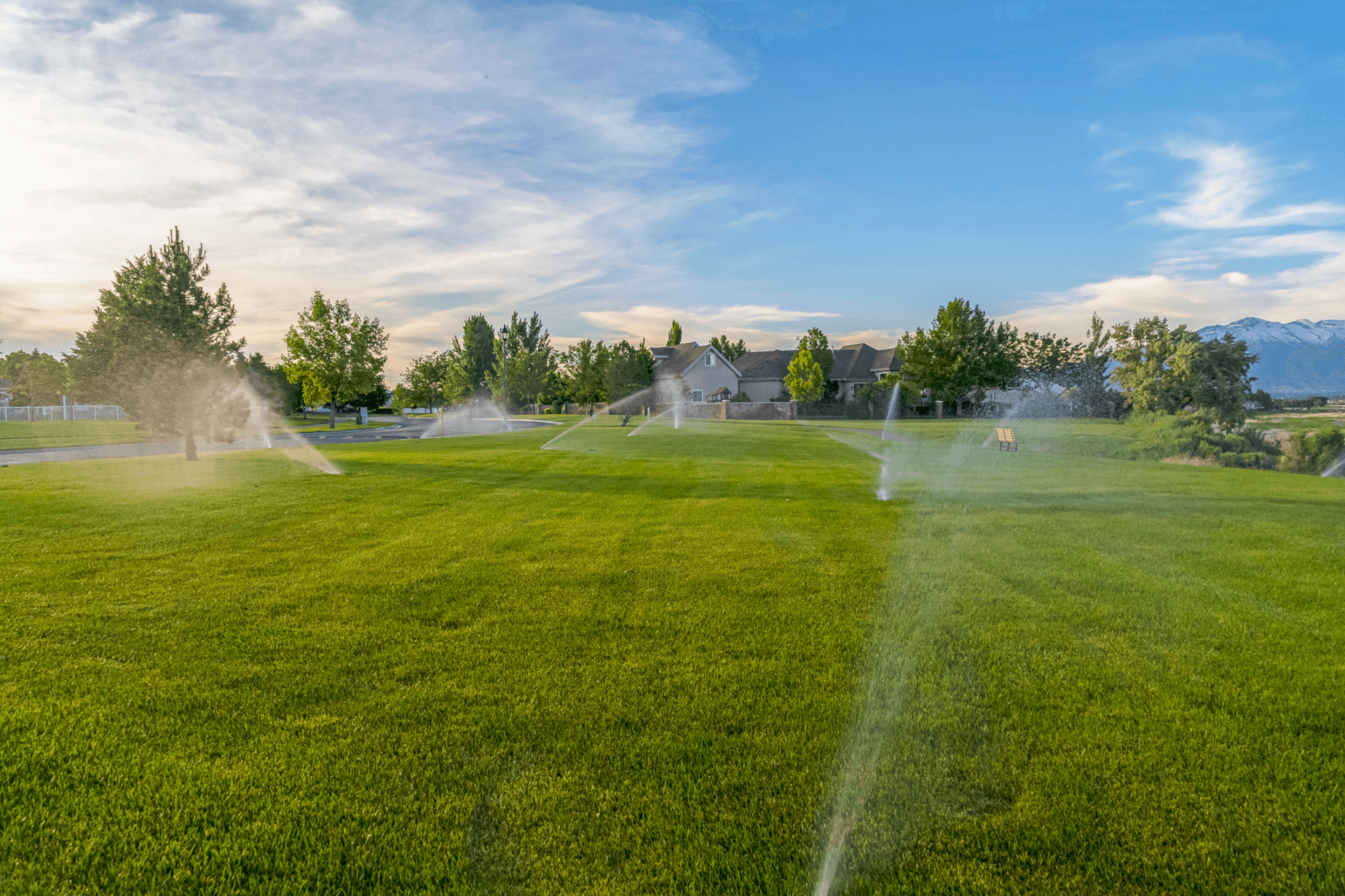 large field of grass being watered by sprinklers in the day with houses and mountains in the background