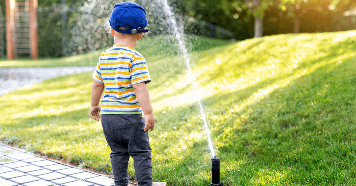 child back faced looking at a single sprinkler near a grass lawn during the day.