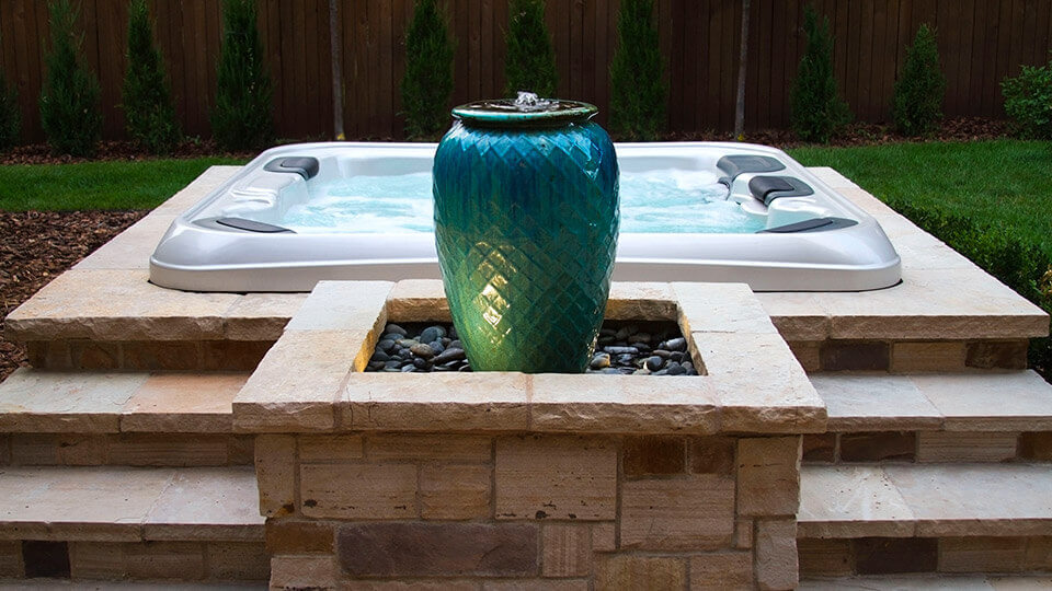 teal fountain on top of a raised stone podium. hot tub behind the fountain with turf grass behind the tub.